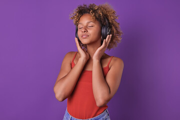 Young optimistic relaxed African American woman teenager in headphones and casual clothes listens to music using wireless technology and enjoying new album of youth singer stands on purple background.