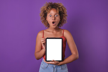 Young shocked pretty African American woman zoomer with electronic tablet in hands opens mouth in admiration and demonstrates white screen of gadget with copy space stands posing in purple studio.