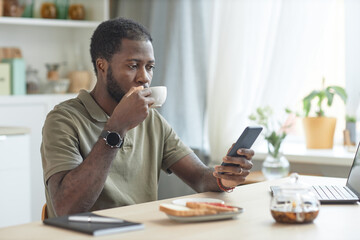 Portrait of adult African American man using smartphone while eating breakfast at kitchen table with laptop