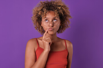 Young pensive curly African American woman touching face remembering forgotten password or coming up with plan for upcoming vacation dressed in red casual clothes stands posing in purple studio.
