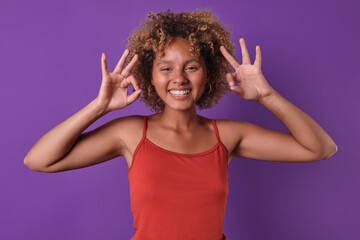 Young admiring ethnic African American woman teenager makes OK gesture with two fingers and shows wide smile when answering question how are you or agreeing to go on date stands on purple background.