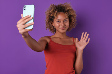Young positive attractive African American woman blogger takes selfie video on mobile phone camera and waves hand greeting subscribers from social networks posing on isolated purple background.