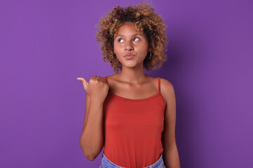 Young pretty shy African American woman teenager looks up and points thumb to side recommending taking advantage of promotional offer to get stands on isolated purple background discount.
