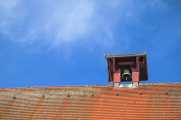 Kleiner Glockenturm mit grüner Glocke auf Ziegeldach vor blauem Himmel mit zarter Wolke in...