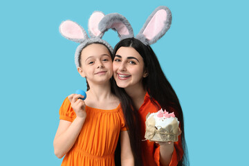 Happy smiling young woman with her daughter in bunny ears holding Easter cake and egg on blue background