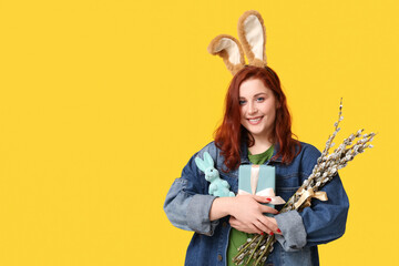 Beautiful young woman in bunny ears with pussy willow branches and gift box on yellow background....
