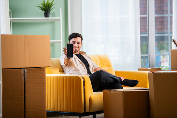 Home moving concept showing indian young man using laptop or smartphone while surrounded with cartons at home