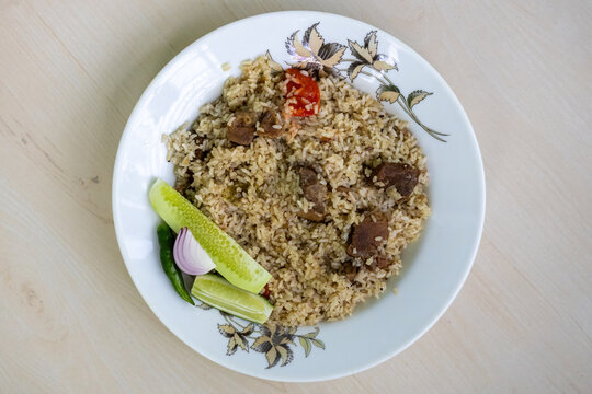 Delicious beef tehari on a white plate with cucumber slices, onion, and green chili on a wooden background. It is a traditional Bangladeshi dish of rice and beef. Top view.