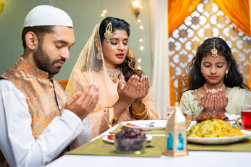 Indian Muslim family with kid praying at ramadan iftar dinner before eating at home - concept of...