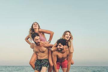 Two couples share laughter with piggyback rides on the beach, embodying the spirit of a carefree...