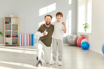 Fototapeta premium Portrait of a happy smiling father standing with his child boy in gym wearing sportswear looking cheerful at camera and showing thumb up sign after workout together. Family sport concept.
