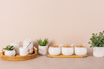 White ceramic tea set on round wicker tray among indoor plants on kitchen countertop. Kitchen background in light colors. A copy space. Front view.