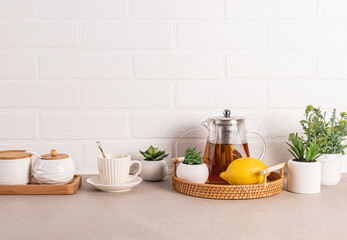 Beautiful kitchen background with glass teapot of fragrant tea, cup, lemon on wicker tray. Front view. White brick wall. A copy space.