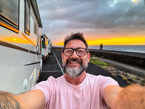 One happy man taking selfie picture on the side of a modern camper van motorhome. Traveler vanlife lifestyle people sharing on social media adventure and journey. Amazing sunset in background. Parking