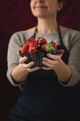 A woman holding a metallic bowl filled with ripe, red strawberries ready to eat, set against a black background