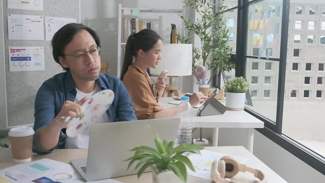 Two Asian workers are unhappy working with hot temperatures at casual office in summer season, uncomfortable with no air conditioner, using fans and drinking water, sitting at desks, and sweating.
