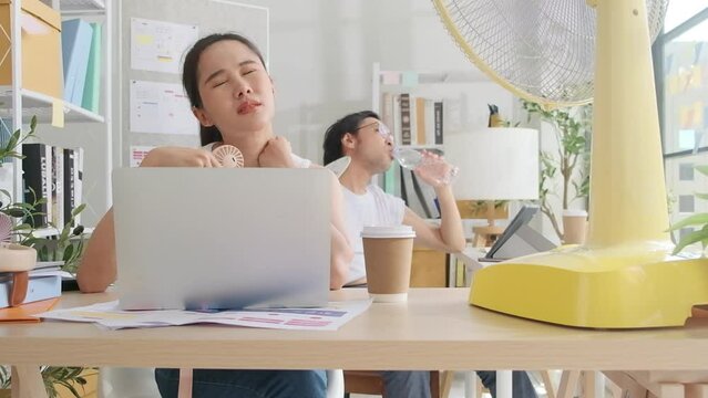 Two Asian workers are unhappy working in hot temperatures at desk in small business office in summer, uncomfortable with no air conditioner, blowing wind with fans, drinking water, and sweating.