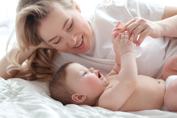 Obraz na płótnie Canvas A 5-month-old baby and his beautiful blonde mom relax, play and laugh in bed in the bedroom. People a dressed in light home clothes, the family chooses natural textiles