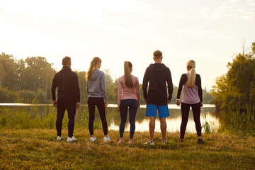 Back view group of people at outdoor fitness workout in nature. View from behind men and women in sportswear standing on beautiful peaceful green lake shore. Sport concept