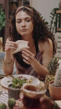 Medium vertical footage of young Mexican woman eating chalupa at dinner table and listening to friends having conversation, while celebrating Cinco de Mayo together