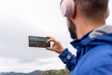 Close-up shot of a man with his back to pink headphones enjoying the music while taking a picture of the coastal landscape with his mobile phone on a cloudy day.