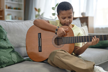 Portrait of young African American boy learning to play acoustic guitar sitting on couch at home...