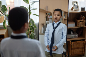 Portrait of young Black boy tying tie and looking in mirror while dressing up at home - obrazy, fototapety, plakaty