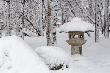 Traditional Japanese stone lantern in a city park. Japanese garden. A lot of snow on the ground, lantern and trees after a snowfall and snowstorm. Cold snowy weather.