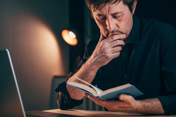 Businessman reading a book while sitting at office desk late at night