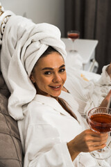 Two young women in bathrobes drink cocktails and pose in the bedroom. Women's party concept