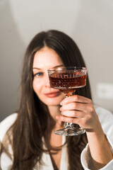 Two young women in bathrobes drink cocktails and pose in the bedroom. Women's party concept