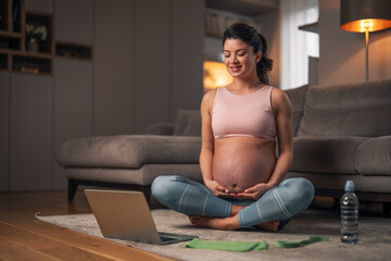 Obraz premium A smiling pregnant lady watching an exercise video on a laptop while holding her belly and sitting on the ground in her apartment