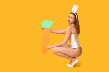 Sexy young woman in bunny ears with paper carrot sitting on yellow background. Easter celebration