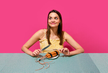 Smiling young woman sitting at table with player and sausages imitating explosive item against pink background. Surrealism. Concept of food pop art photography, creativity, quirky style