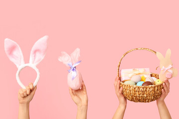 Female hands holding basket with Easter eggs, bunny ears and gift on pink background