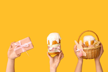 Female hands holding Easter cake with basket and gift box on yellow background