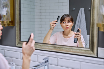 Middle-aged woman with setting spray for styling hair fixation comb looking in mirror