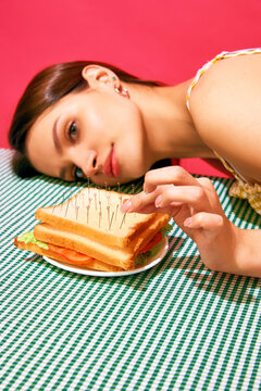Young woman lying next to sandwich with pins resembling acupuncture against pink background. Meditation. Concept of food pop art photography, creativity, quirky style