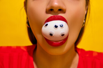 Close-up of woman's face with red lipstick, egg on mouth with googly eyes against yellow background. Concept of food pop art photography, creativity, quirky style