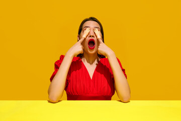 Woman in red dress with surprised expression, covering eyes with toy hands against yellow background. Emotive woman. Concept of food pop art photography, creativity, quirky style