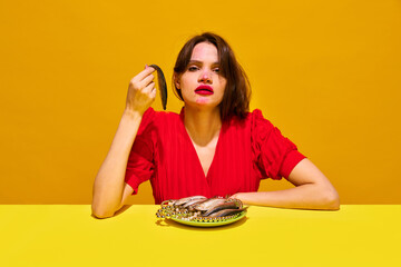 Young woman with smudged lipstick makeup eating fish against yellow background. Beauty standards and societal consumption. Concept of food pop art photography, creativity, quirky style