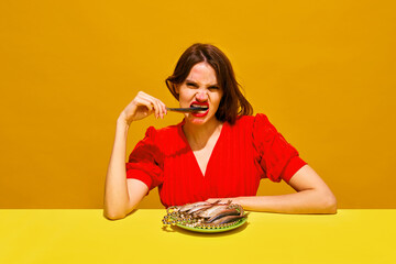Young woman with smudged lipstick makeup eating fish against yellow background. Expressive dining and food culture. Concept of food pop art photography, creativity, quirky style
