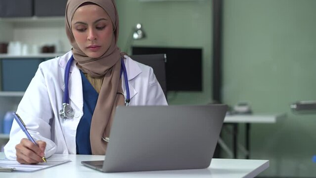 Female medicine doctor, physician or practitioner in lab room writing on blank notebook and work on laptop computer with medical stethoscope on the desk at hospital. Medic tech concept.