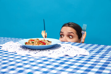 Young woman peeking over table, gazing at plate of noodles with toy hand holding knife and fork against blue background. Concept of food pop art photography, creativity, quirky style
