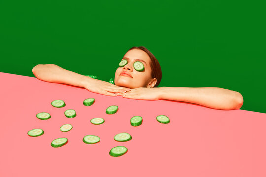 Spa and relaxation. Young woman peeking over table with cucumber slices on face, homemade refreshing mask against green background. Concept of food pop art photography, creativity, quirky style