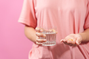 Graciously, woman hand holds  bottle of cod liver oil and a glass of water against a pink backdrop, illustrating health, hydration, and nutritional supplementation.