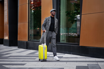 Stylishly dressed male model walks down city street with yellow suitcase in hand. Black man tourist outdoors near hotel building. African american man in trendy coat, cap and Luggage