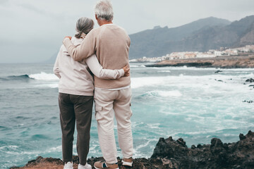 Rear view of a romantic senior couple embrace on the seashore on a bad weather day looking waves...