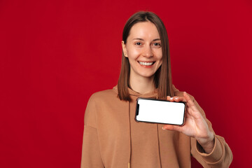 Excited young woman is holding a phone with blank screen in a studio with red background.