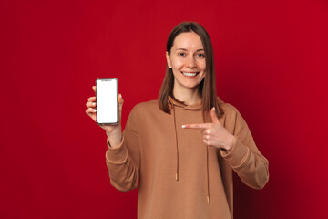 Cheerful young woman is pointing at the phone with blank screen she is holding. Studio shot over red background.